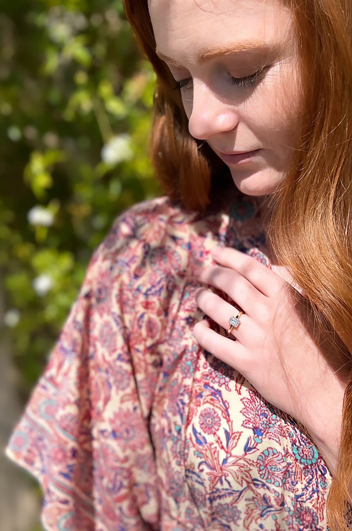 Kate with her hand across her chest showing her new engagement ring, a simple yellow gold solitaire ring featuring a prong set emerald cut diamond 