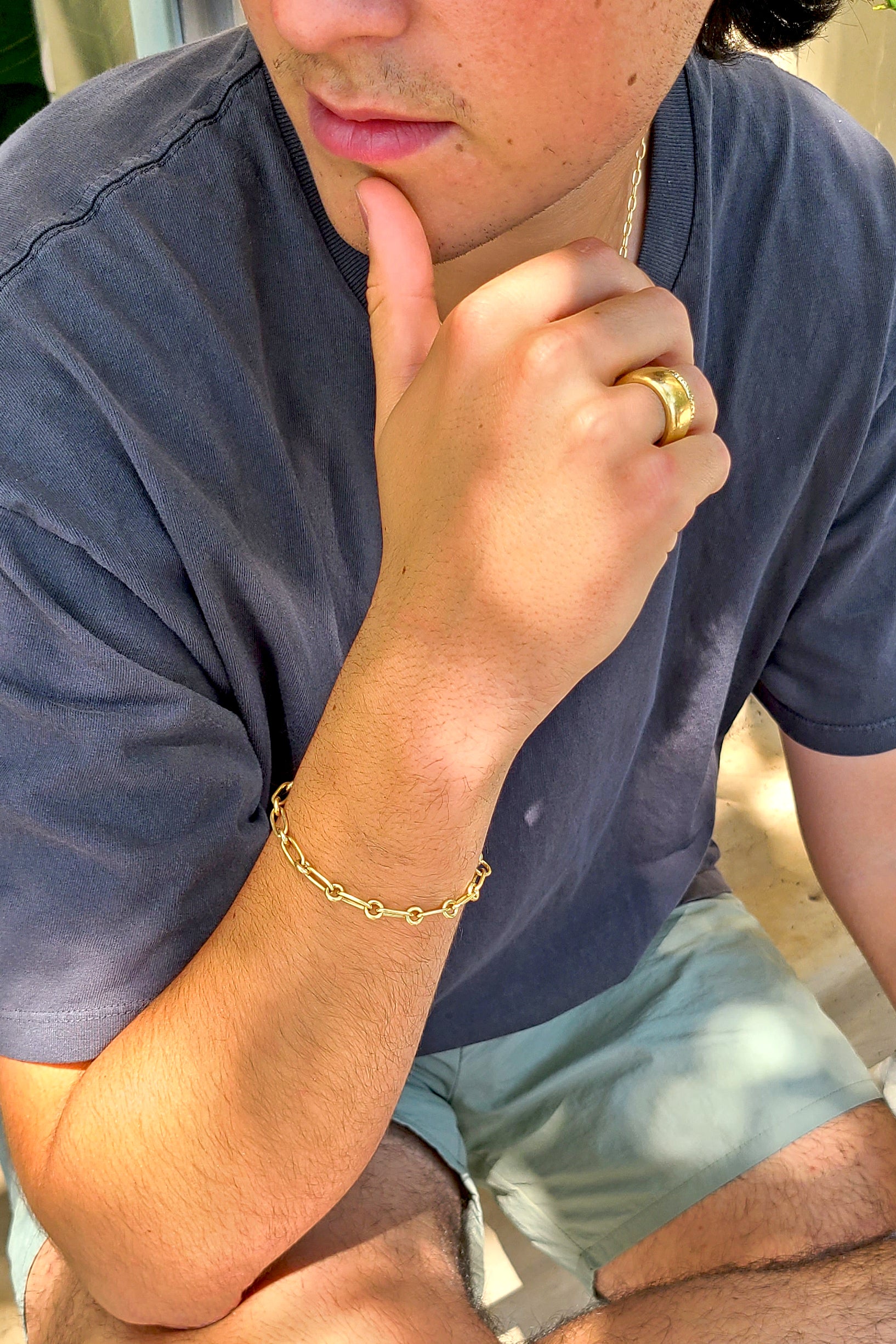 man in a gray shirt wearing a gold chain link bracelet and two textured gold bands.