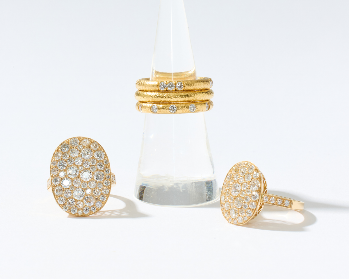Three gold rings displayed on a clear cone stand against a clean white background. Two oval pavé rings sit on either side of the stand, and a stack of three slim gold rings with small clear stones is centered on the stand.