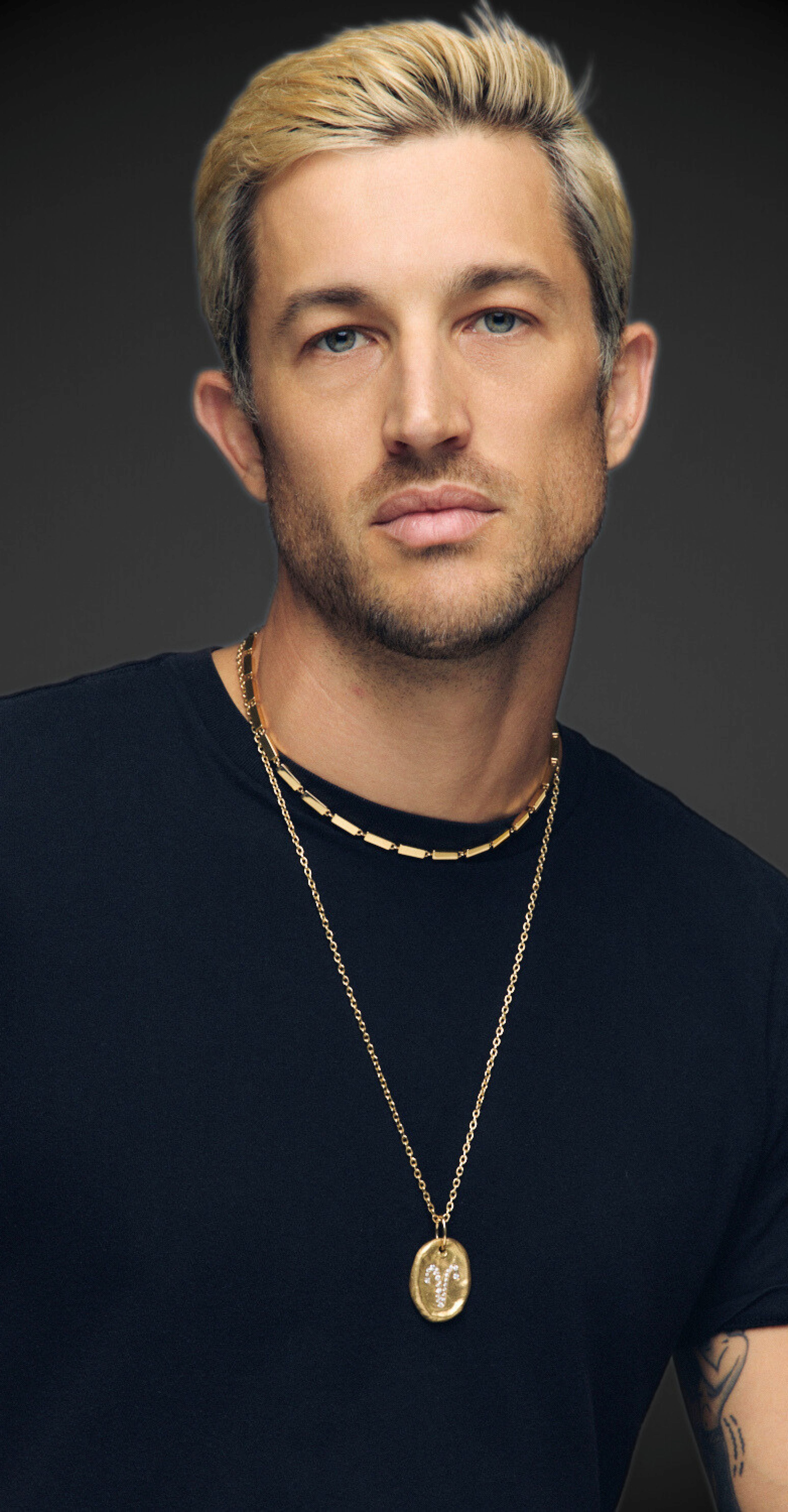 Man wearing a black shirt and gold necklace with a pendant against a dark background