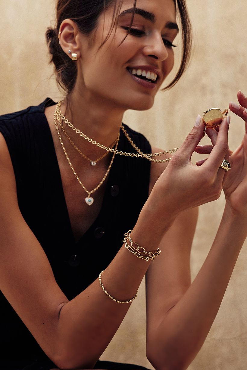 Model holding up an engravable vintage-style locket in yellow gold, with its chain around her neck, and smiling. Other Single Stone vintage-inspired necklaces are layered around her neck.