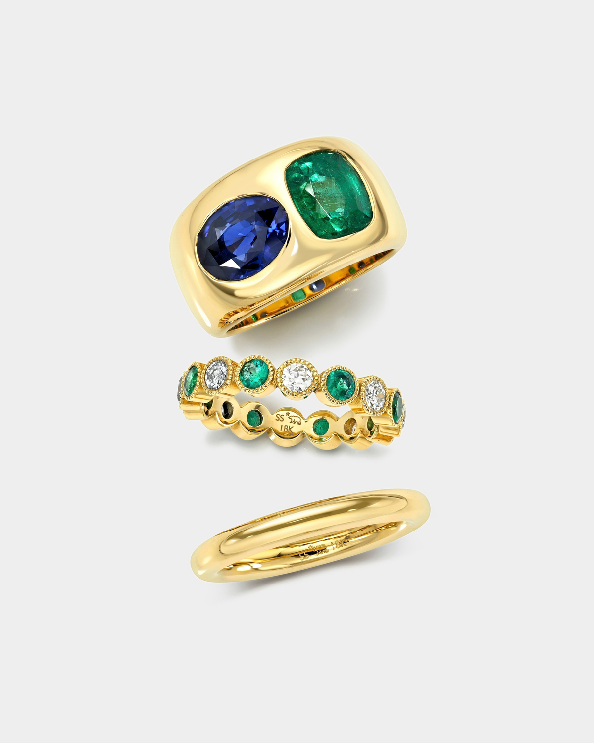 Three gold rings displayed on a light gray background: a wide ring with a large oval blue sapphire and a rectangular emerald-cut green gemstone, a delicate eternity-style band with alternating green and clear stones, and a slim polished gold band.
