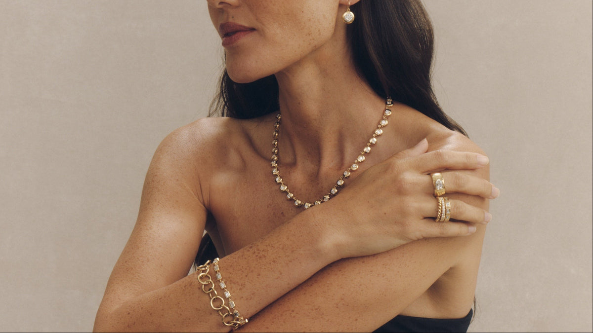 This image shows a woman with long dark hair, wearing a black top, and adorned with several pieces of jewelry. She is wearing a necklace with a series of small, round, golden beads. On her left ear, she has a small, round, golden earring.