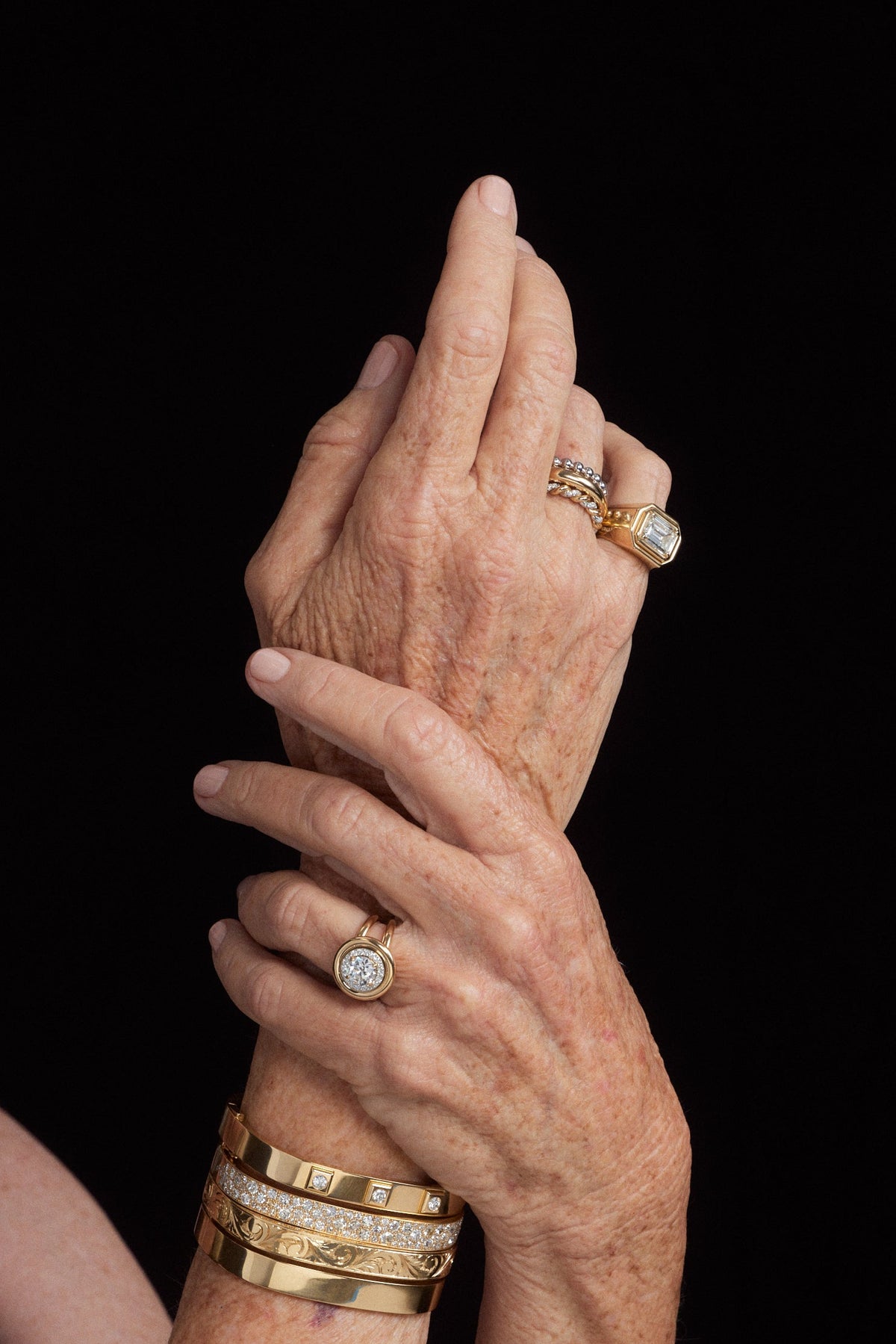 Close-up of crossed hands wearing multiple gold jewelry pieces, including rings and stacked bracelets, against a solid black background.