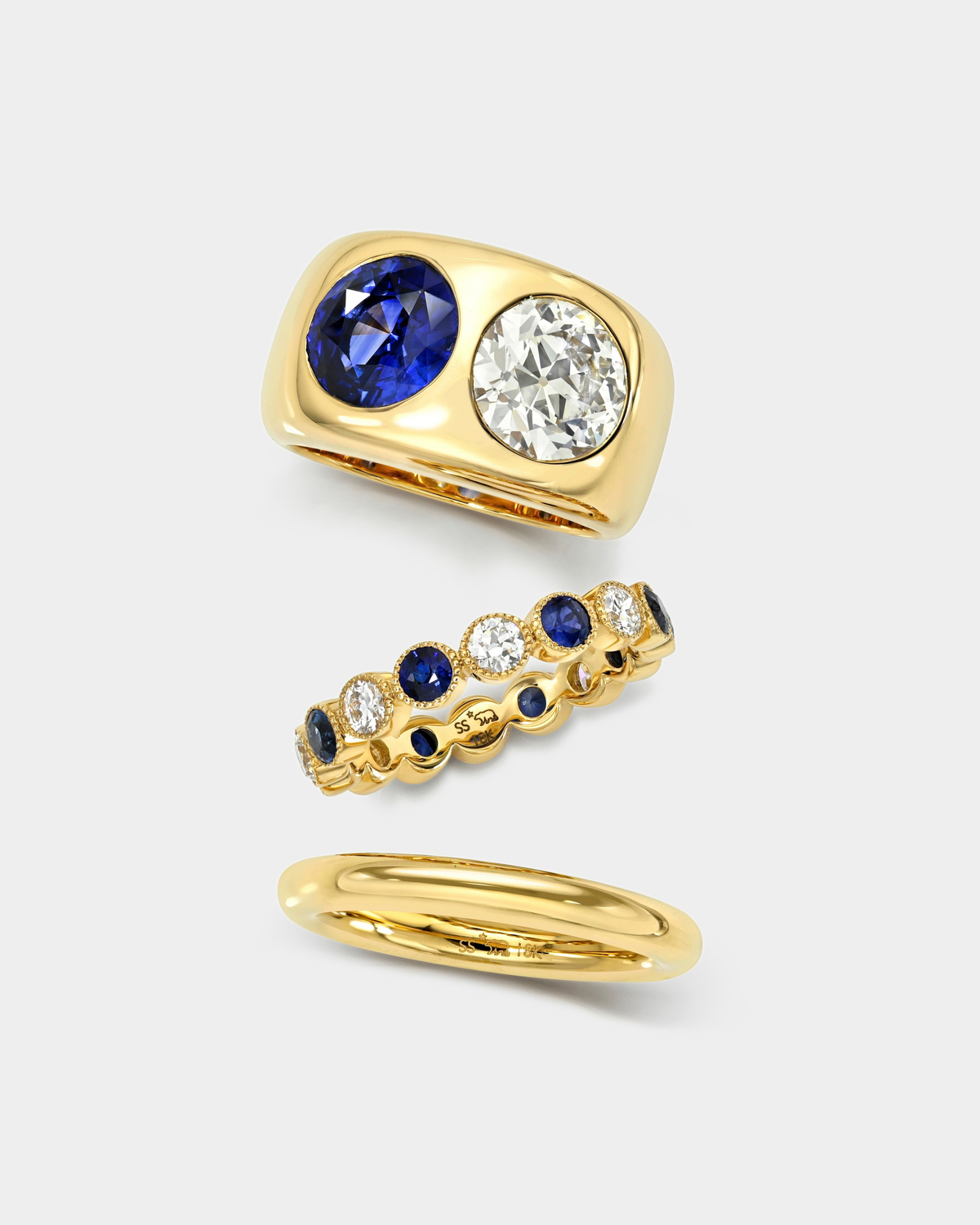 Three yellow gold rings displayed on a light gray background: a wide band ring with a large oval blue sapphire and a round clear diamond, a slim eternity-style band ring with alternating blue and clear stones, and a simple polished gold band.