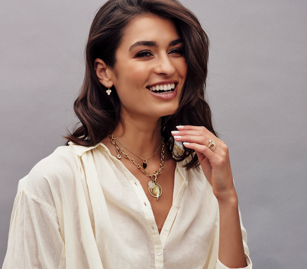Woman wearing layered gold necklaces, rings and bands, and diamond drop earrings from the Single Stone collection. She is holding her hand close to her mouth and laughing.