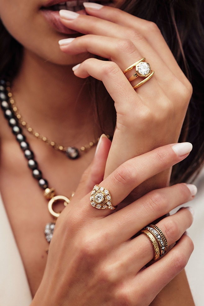 Close up of a woman's hands wearing several gold rings and bands from the Single Stone collection. She is holding her hands up close to her mouth and the Tahitian pearl necklace she's wearing is visible in the background.