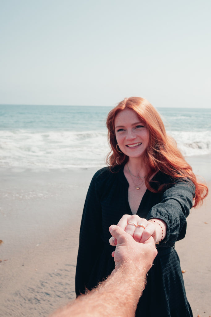Kate smiling, her hand outstretched showing her engagement ring, clasped with Tommy's hand who is otherwise off camera, with sand and ocean as the backdrop