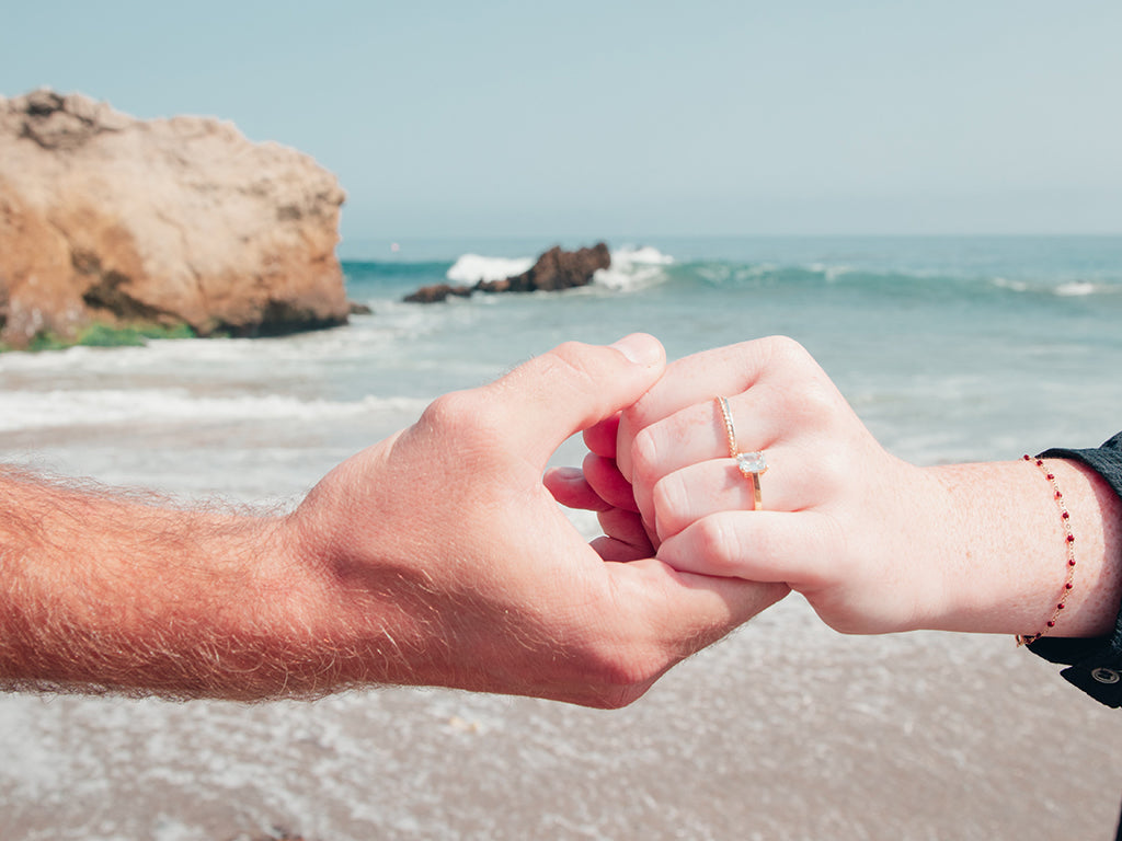 A woman's hand wearing a diamond engagement ring, clasped with a man's hand with the ocean as the backdrop