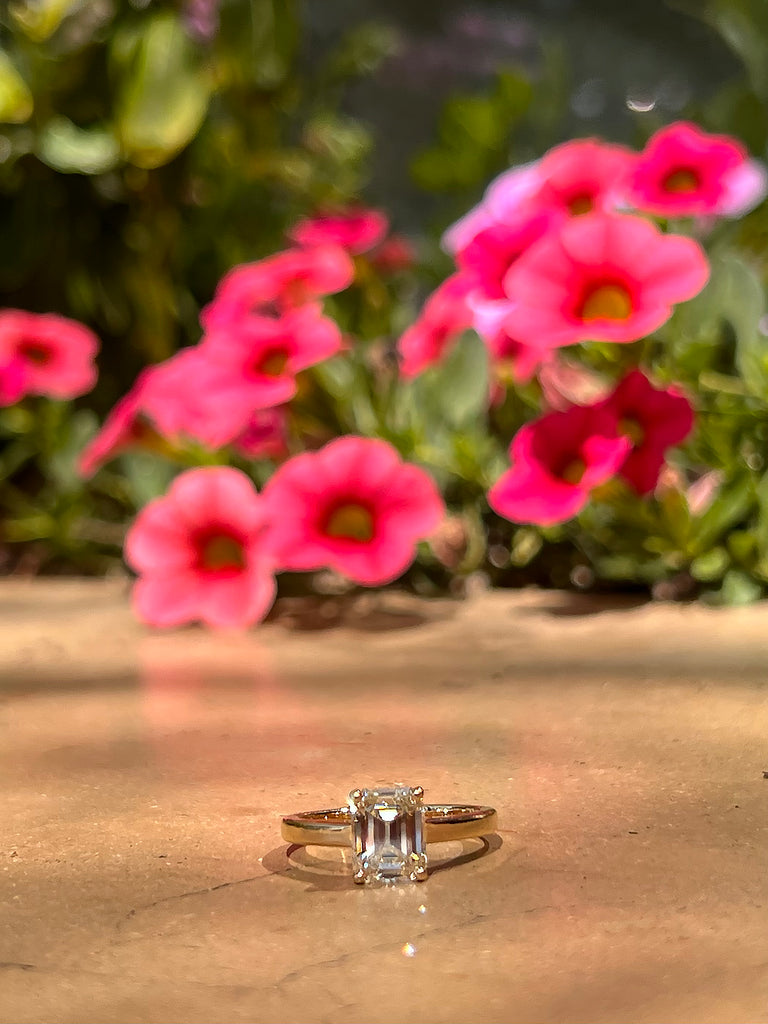 Kate's engagement ring, a simple yellow gold solitaire ring featuring a prong set emerald cut diamond, with pink and green flowers in the background