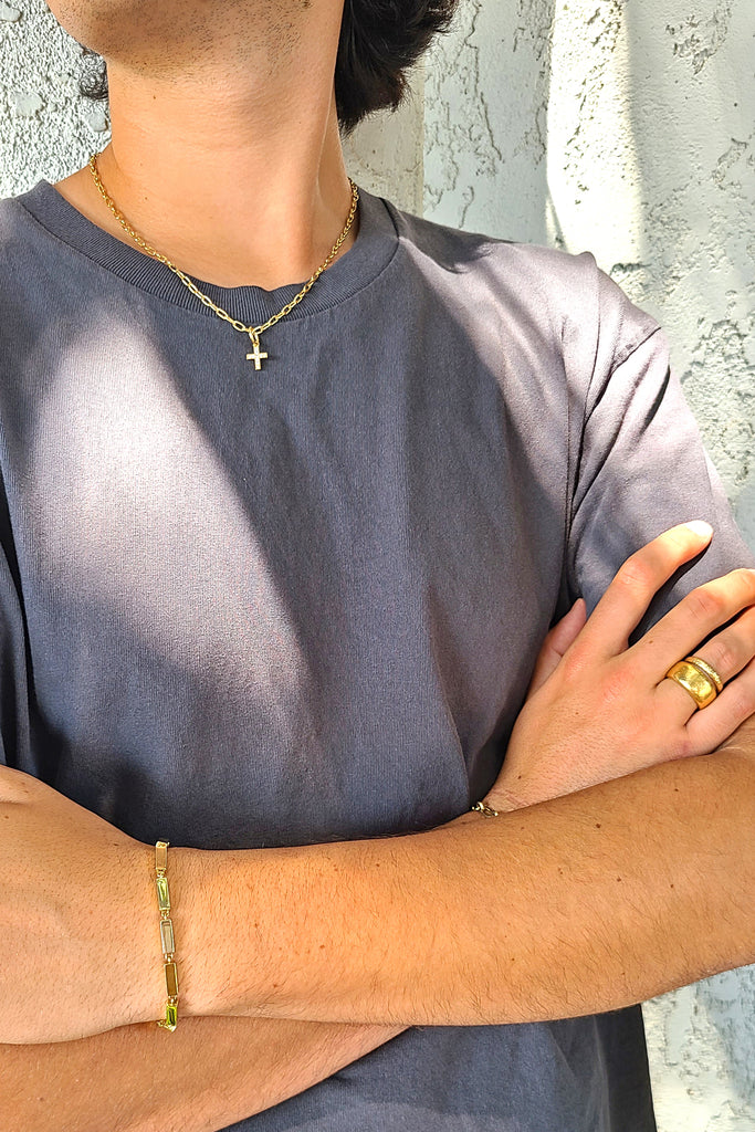 man in a gray shirt wearing a gold chain link necklace with a cross-shaped diamond pendant, a gold bar bracelet, and two textured gold bands