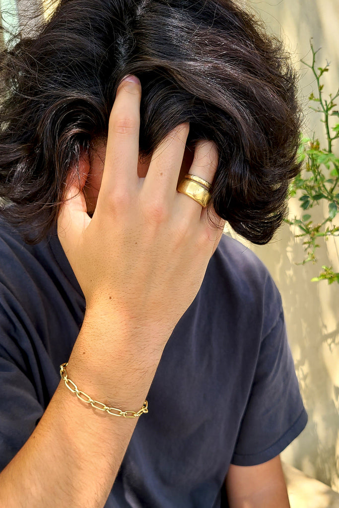 man in a gray shirt wearing a gold chain link bracelet and two textured gold bands.