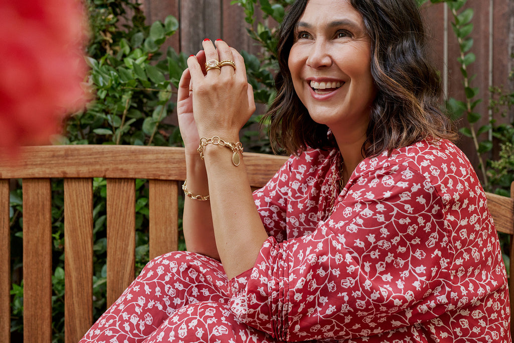 Single Stone Designer Corina Madilian sitting on a bench and smiling in a red and white floral dress and Single Stone jewelry