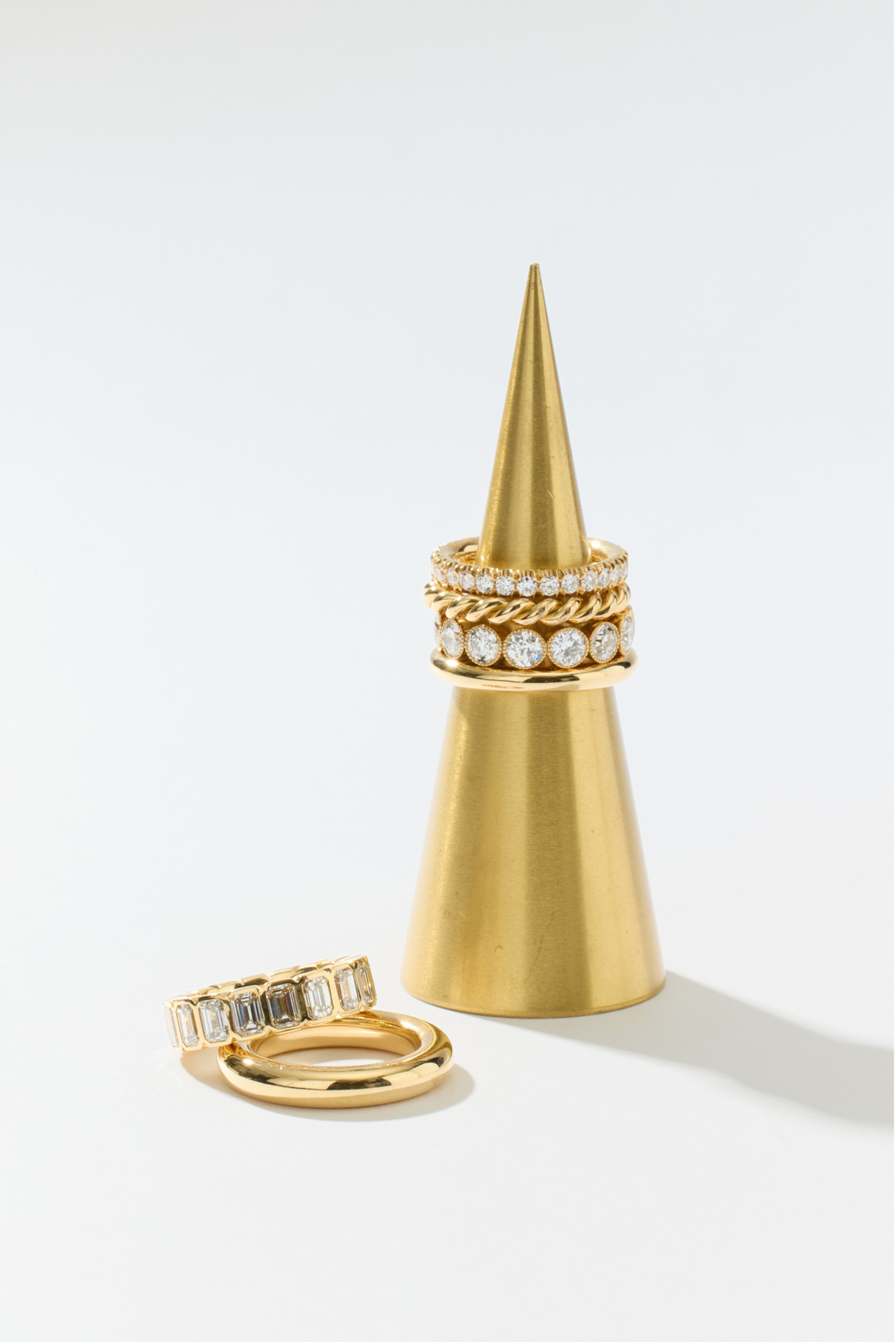 A set of gold rings displayed on a gold cone ring stand against a clean white background. One ring is shown standing on its side, and another set of rings is stacked on the cone, featuring clear gemstones.