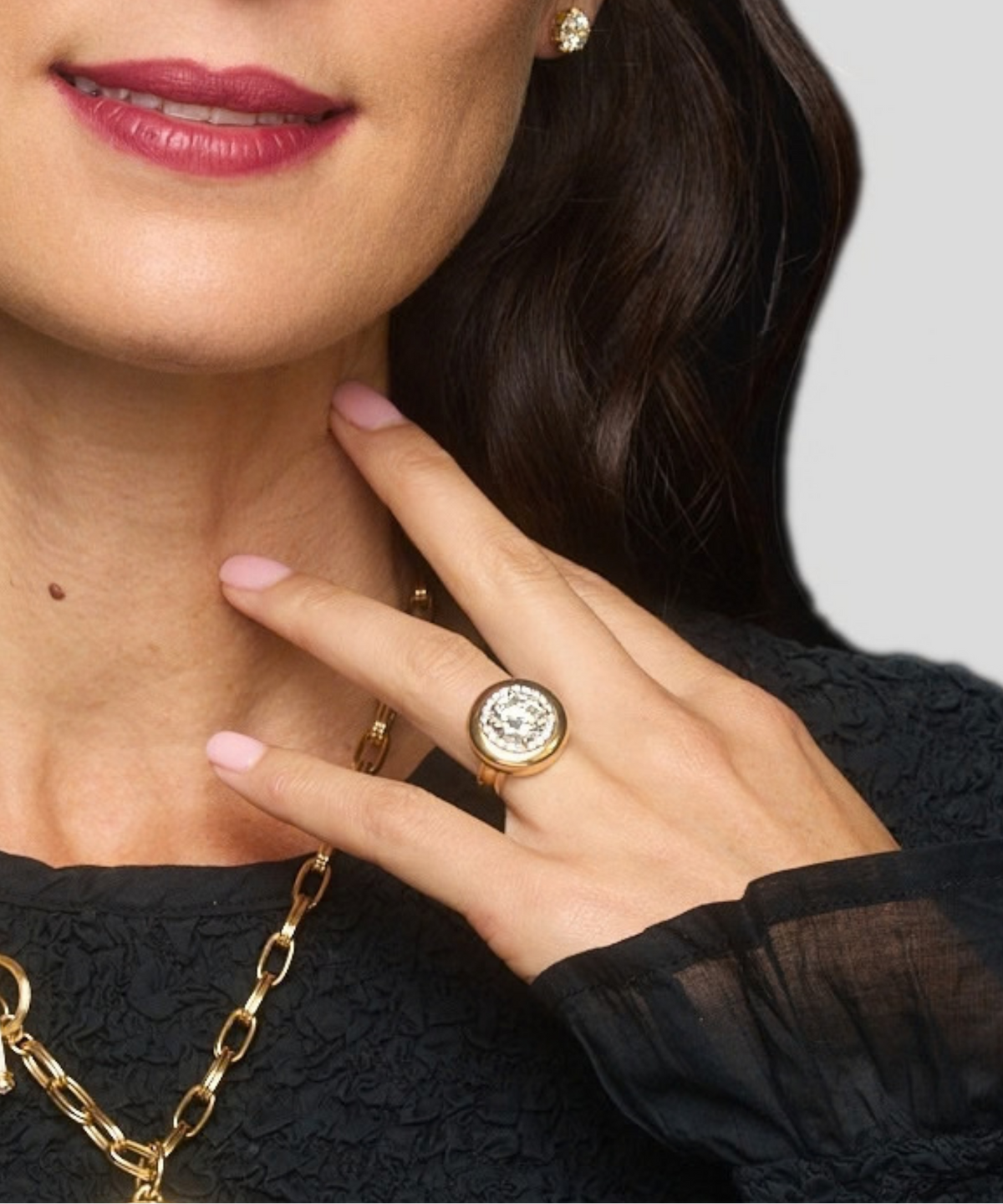 A close-up lifestyle shot of a model wearing a gold signet ring with a clear round gemstone. The model is wearing a black lace top, a gold chain necklace, and a small gold stud earring against a light gray background.