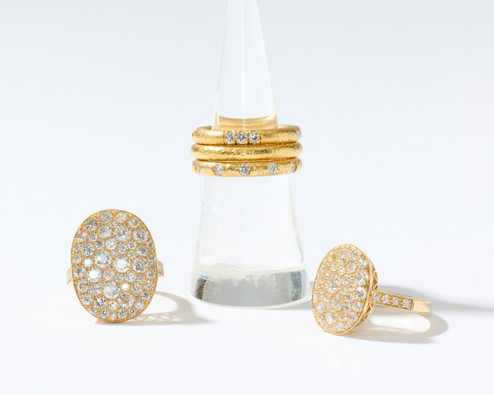 Three gold rings displayed on a clear cone stand against a clean white background. Two oval pavé rings sit on either side of the stand, and a stack of three slim gold rings with small clear stones is centered on the stand.