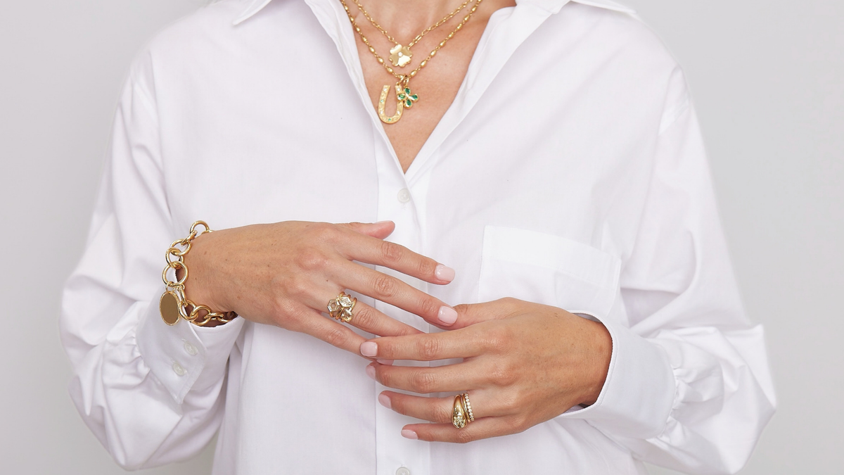 A close-up of a person wearing a white button-up shirt and layered gold jewelry, including necklaces, bracelets, and rings, against a light gray background.