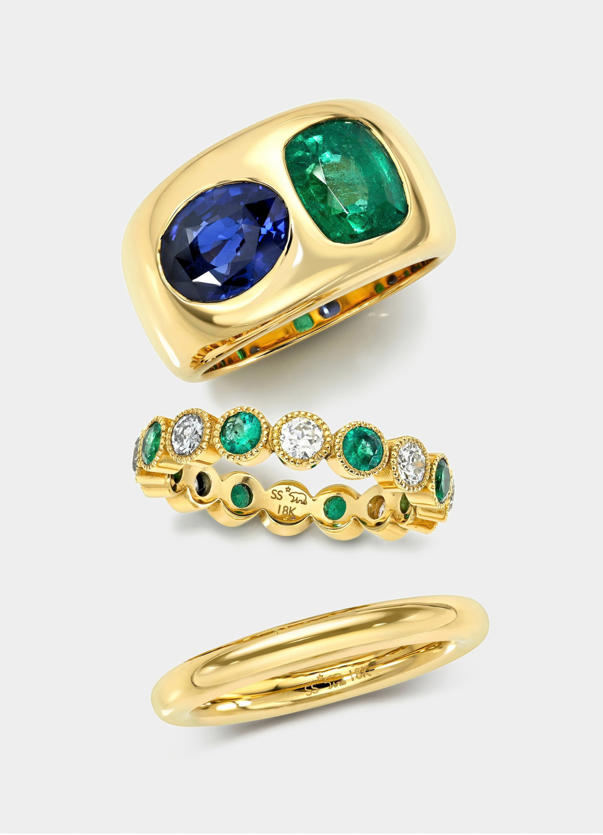 Three gold rings displayed on a light gray background: a wide ring with a large oval blue sapphire and a rectangular emerald-cut green gemstone, a delicate eternity-style band with alternating green and clear stones, and a slim polished gold band.