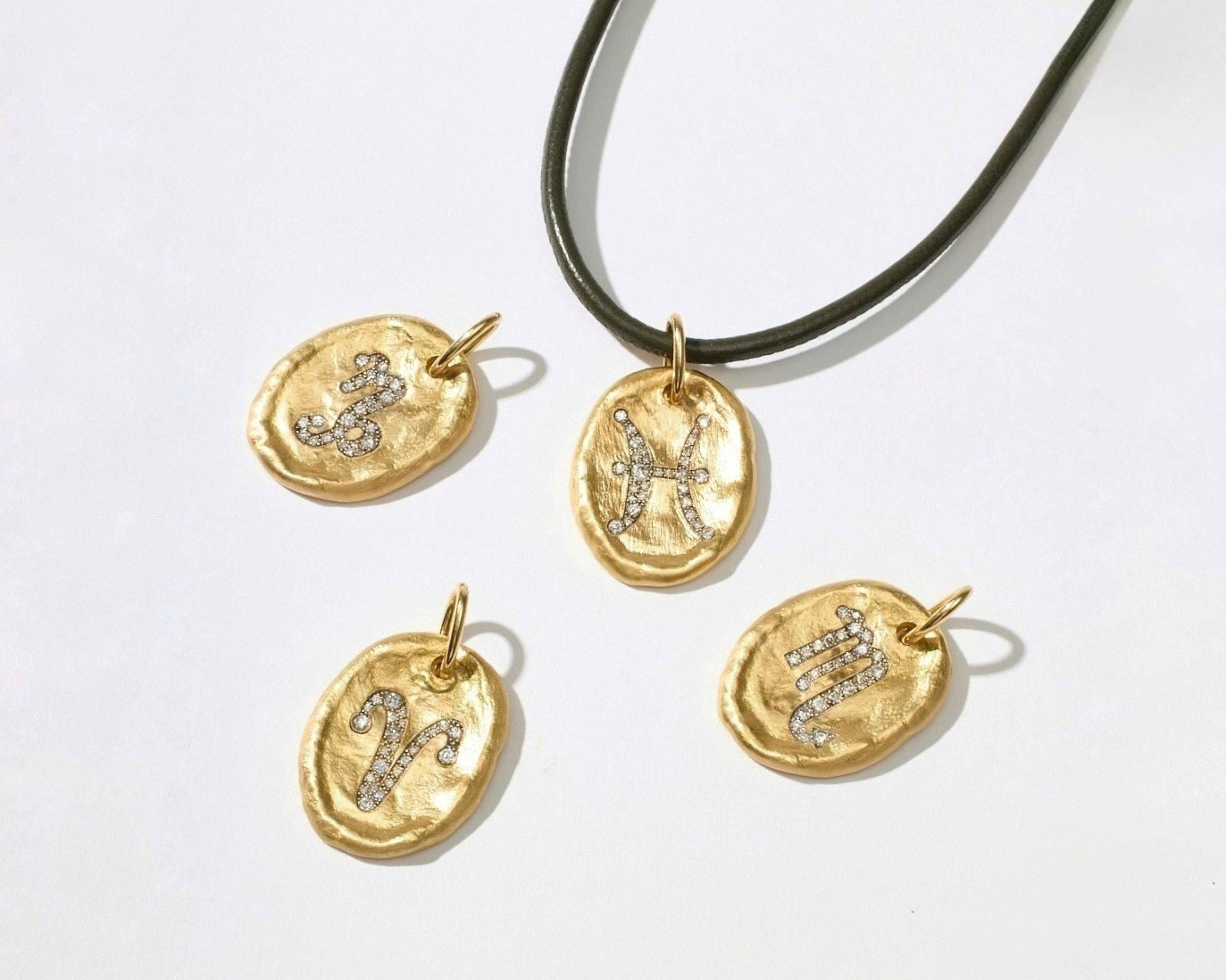 Four gold-toned oval pendant charms with engraved zodiac symbols, some set with small clear stones, displayed on a white background with one charm hanging from a black cord.
