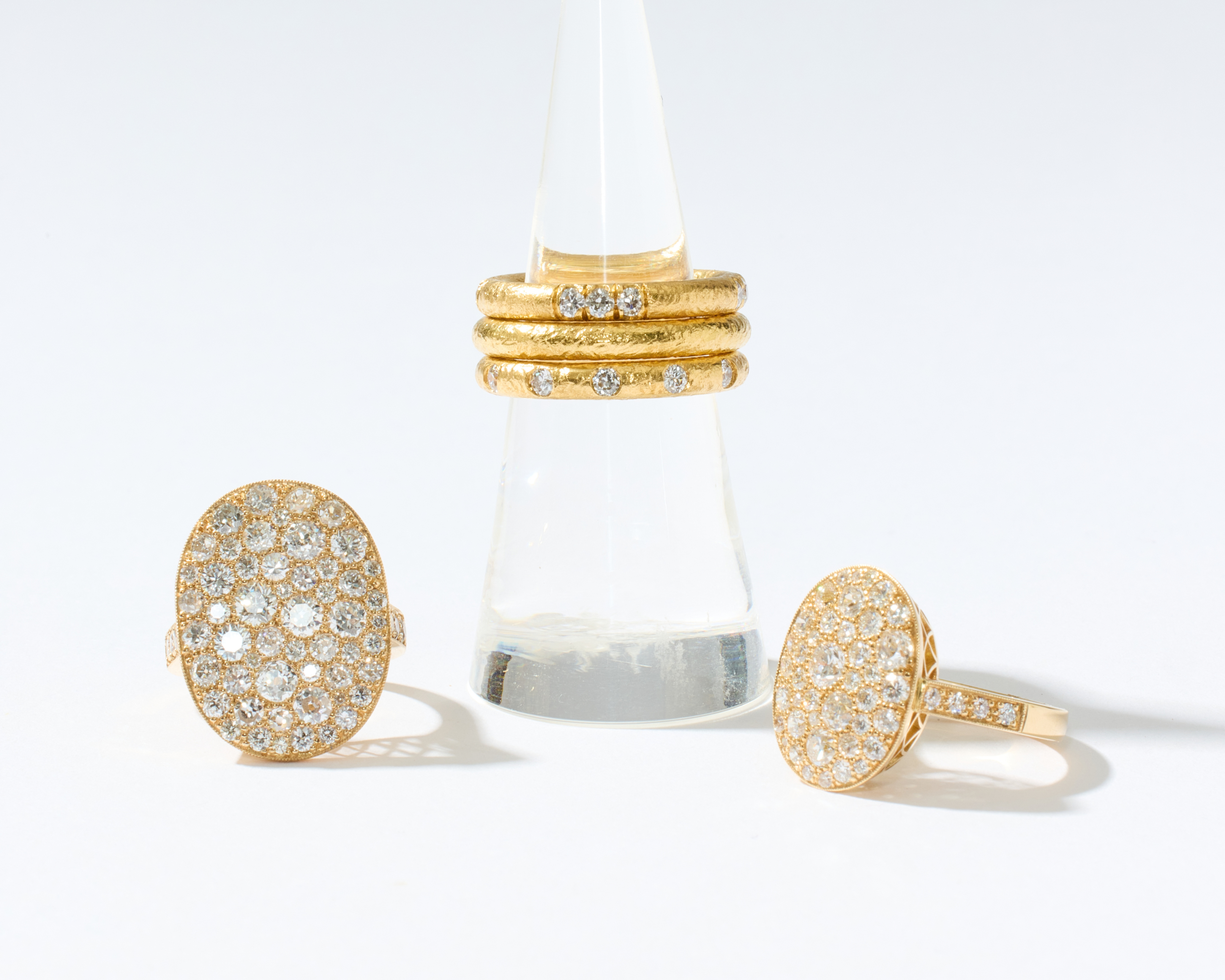 Three gold rings displayed on a clear cone stand against a clean white background. Two oval pavé rings sit on either side of the stand, and a stack of three slim gold rings with small clear stones is centered on the stand.