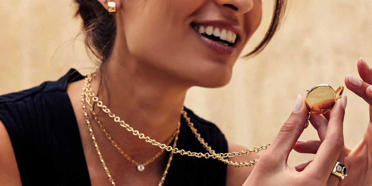 Model holding up an engravable vintage-style locket in yellow gold, with its chain around her neck, and smiling. Other Single Stone vintage-inspired necklaces are layered around her neck.