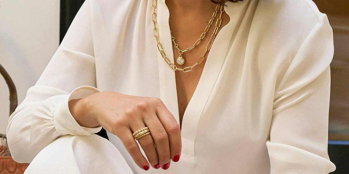 A close-up of a person wearing layered gold necklaces and a gold ring, dressed in a white blouse.