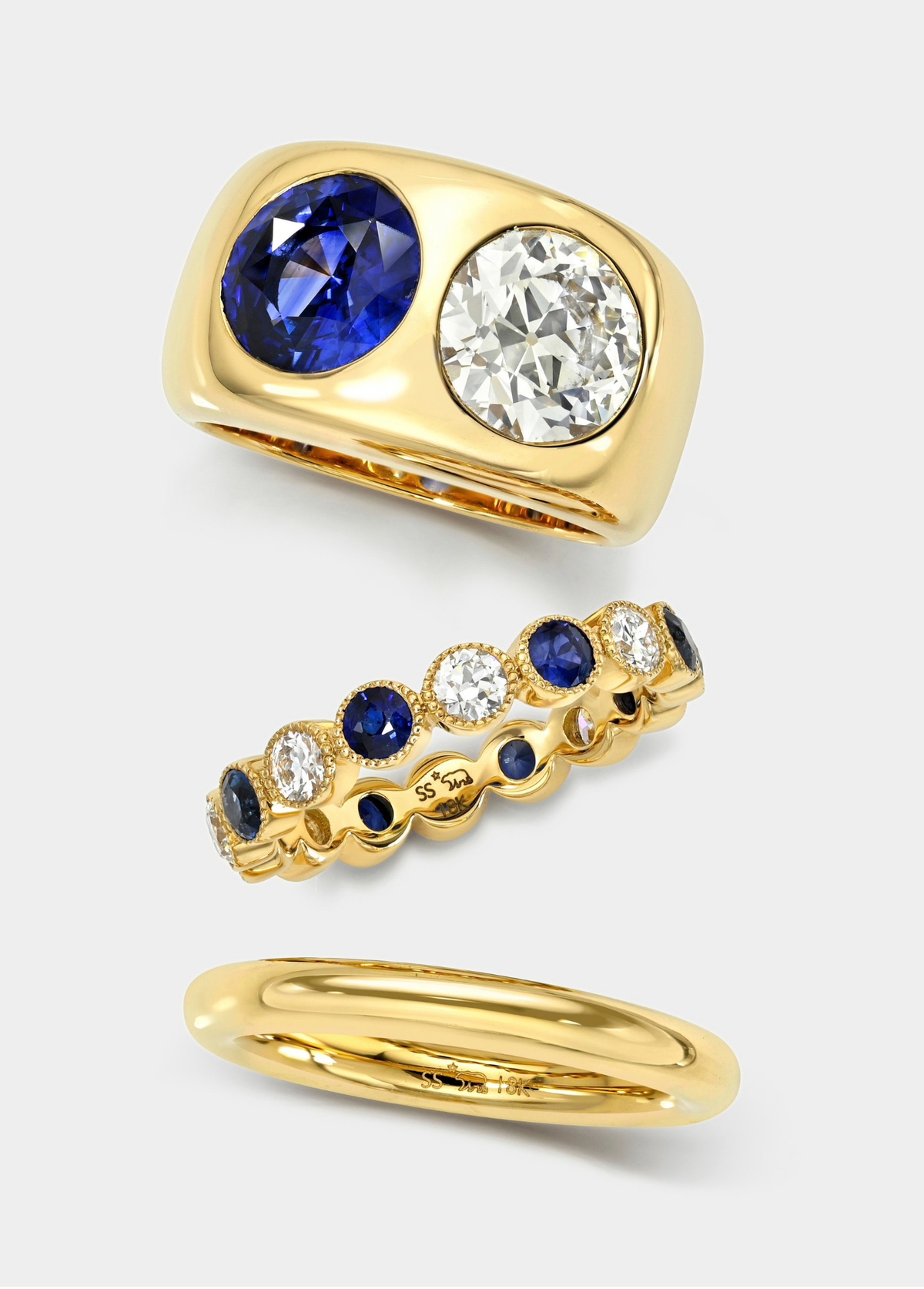 Three yellow gold rings displayed on a light gray background: a wide band ring with a large oval blue sapphire and a round clear diamond, a slim eternity-style band ring with alternating blue and clear stones, and a simple polished gold band.