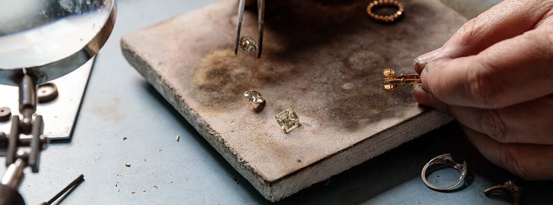 Antique diamonds on a work surface with tweezers holding one antique diamond just above the surface and a hand holding an empty ring setting.