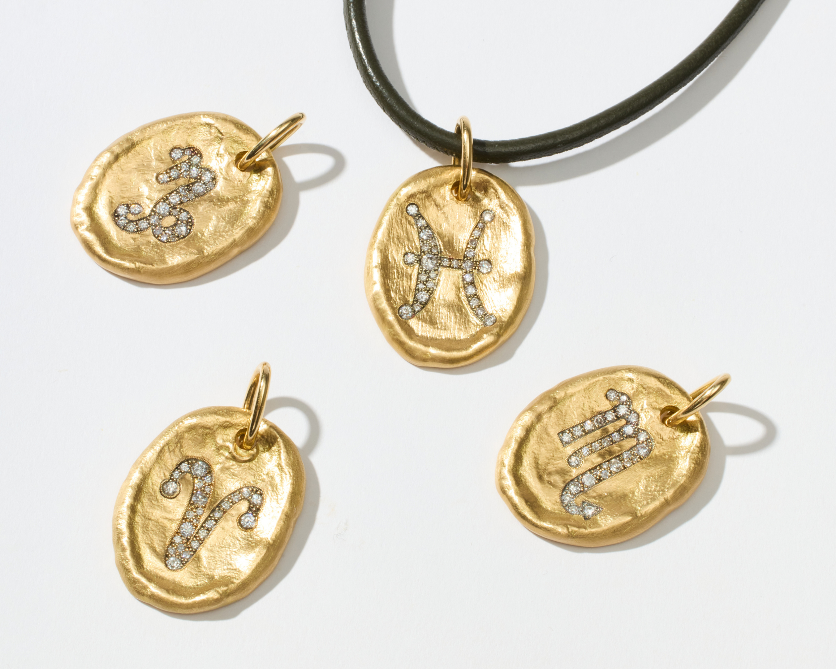 Four gold-toned oval pendant charms with engraved zodiac symbols, some set with small clear stones, displayed on a white background with one charm hanging from a black cord.