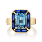 SINGLE STONE PIPPA RING featuring 2.63ct emerald cut aqua surrounded by 1.25ctw French cut blue sapphire baguette accent stones set in a handcrafted 18K yellow gold mounting.